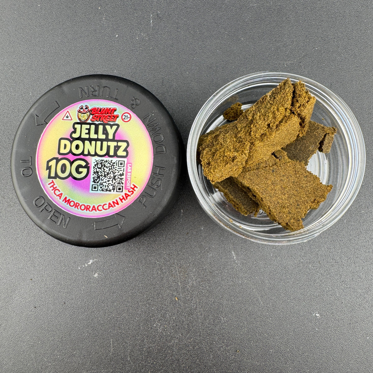 A 10g roll of Jelly Donutz THC Mororacan Mesh disposable joints shown next to its clear plastic container filled with brown, textured, crumbly pieces of the product, on a dark gray background.
