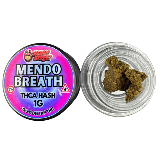 A small clear plastic cup containing dark green-brown dried plant material sits below a round black-lidded container labeled 'MENDO BREATHE 1G' with a colorful pink, purple, and blue label. The product is photographed on a dark gray textured surface.