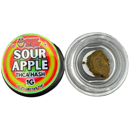 A small clear jar filled with dark brown-green THC hash next to its lid, which has a colorful label reading 'Sour Apple' and 'Delta Pine'.