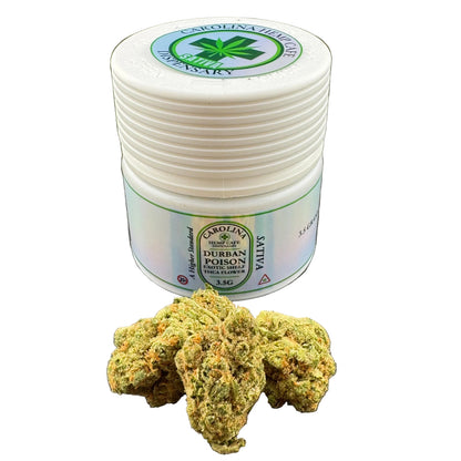 A white plastic jar of Carolina Herbs Apothecary Durban Poison Sativa flower with a green and white label, shown next to several green and orange trichome-covered cannabis buds on a dark gray surface.