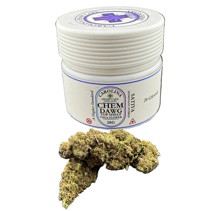 A white plastic jar of Carolina HempCare Chem Dawg Top Shelf Sativa flower with a green and brown cannabis bud placed in front of it on a dark gray background.