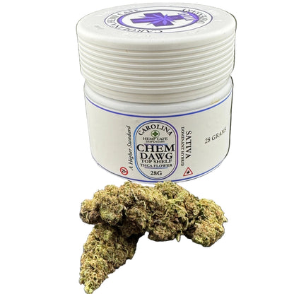 A small white plastic jar labeled Carolina Top Shelf Chem Dawg Sativa 28g, shown on a dark gray surface with a cannabis flower bud placed in front of it.