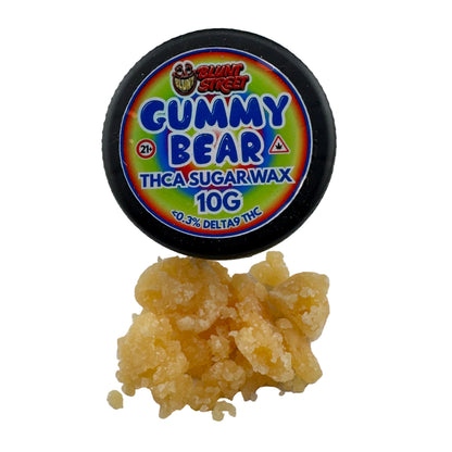 A 10g tub of Gummy Bear THCA Sugarwax with the lid propped open, showing a pile of translucent golden-yellow wax crystals below on a white background.