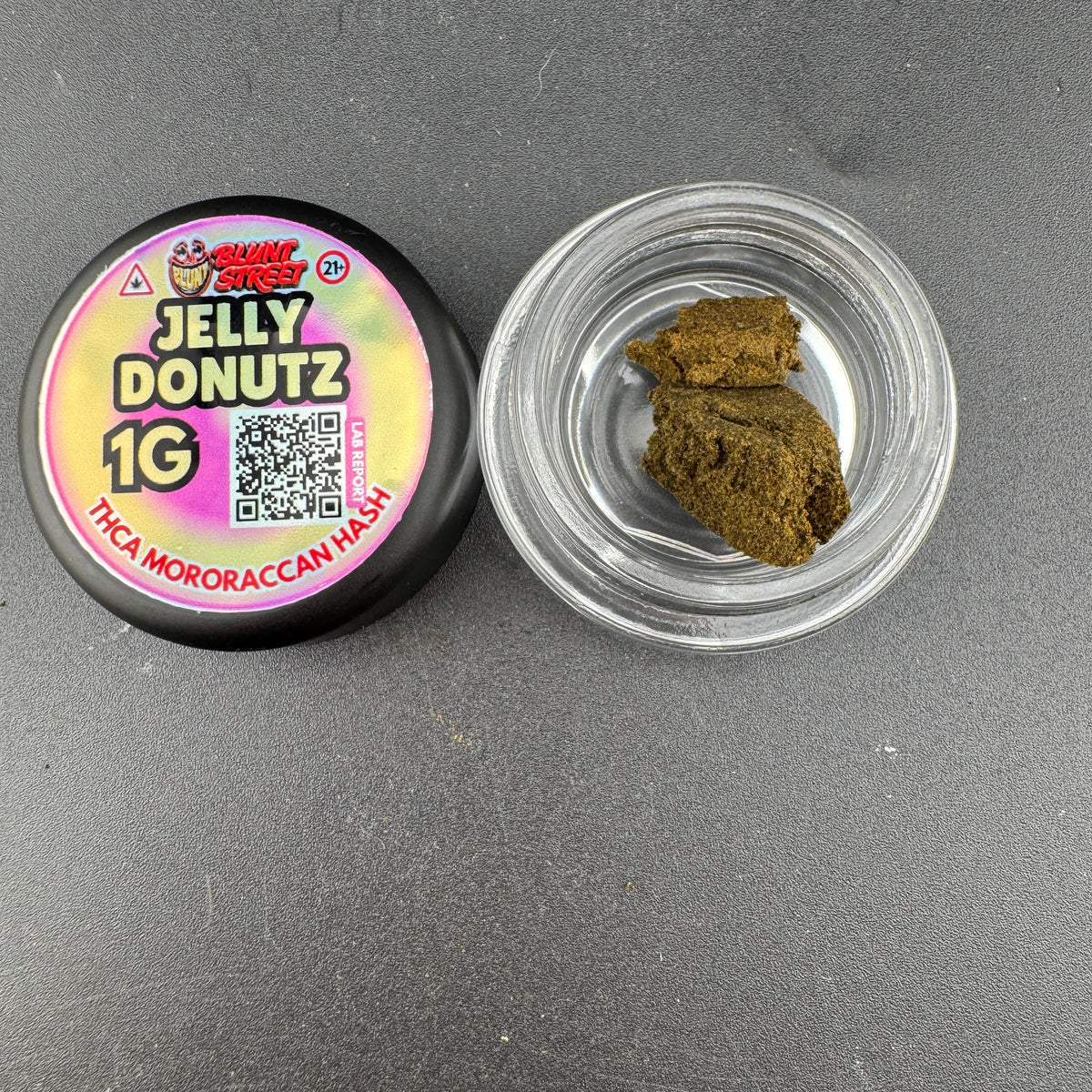 A 1g jar of Jelly Donutz THCa Mororaccan Hash by Shady Stacks, showing the black lid with a colorful label next to an open clear jar containing brownish-green hash.