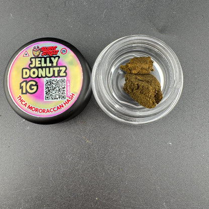 A 1g jar of Jelly Donutz THCa Mororaccan Hash by Shady Stacks, showing the black lid with a colorful label next to an open clear jar containing brownish-green hash.